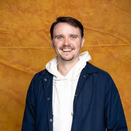 Comedian Pete Ballmer smiling against a warm gold backdrop