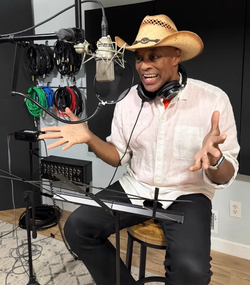 Robby the Vibe in a straw cowboy hat and headphones, talking animatedly at a studio microphone